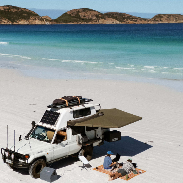 Aerial view of a Troop Carrier on a white-sand beach, its 270 XT Awning Mk2 shading a couple on a rug