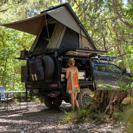 Woman beside a canopied ute with the AX27 rooftop tent open, at a campsite among trees