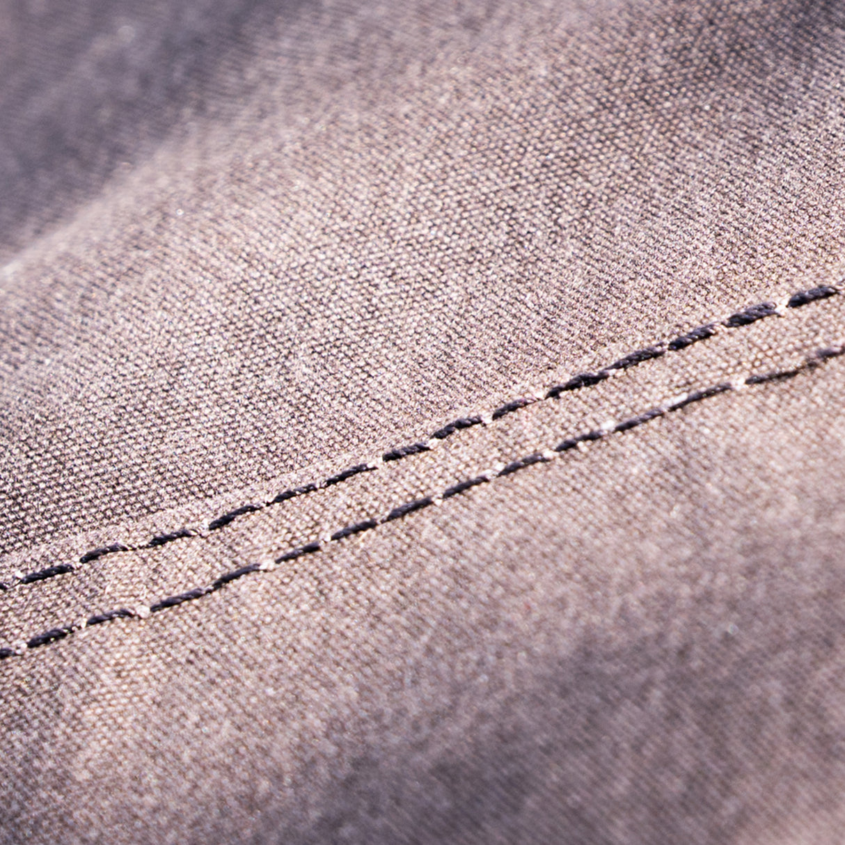 Close-up of a woven fabric surface in a muted taupe-brown colour with a visible double-stitched seam running diagonally across the frame.