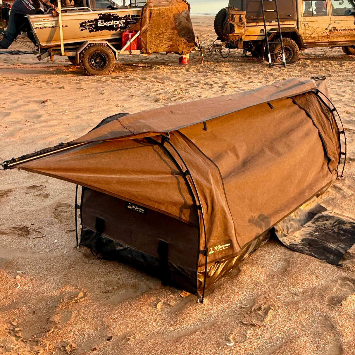 A brown canvas camping swag with a black base and black frame, set up on sandy ground with off-road vehicles in the background.
