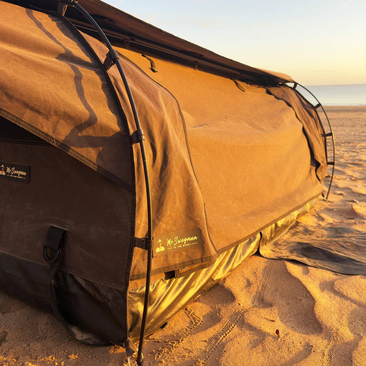 A brown camping swag pitched on a sandy beach during golden hour, with the ocean and sky visible in the background.
