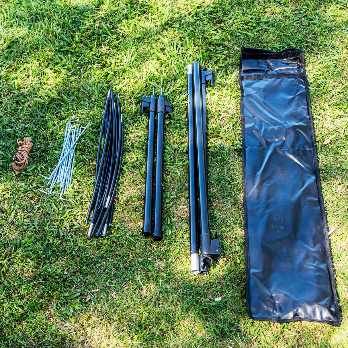 A black waterproof tent body laid out on green grass alongside its black poles, silver stakes, and a brown roll of waterproof tape.