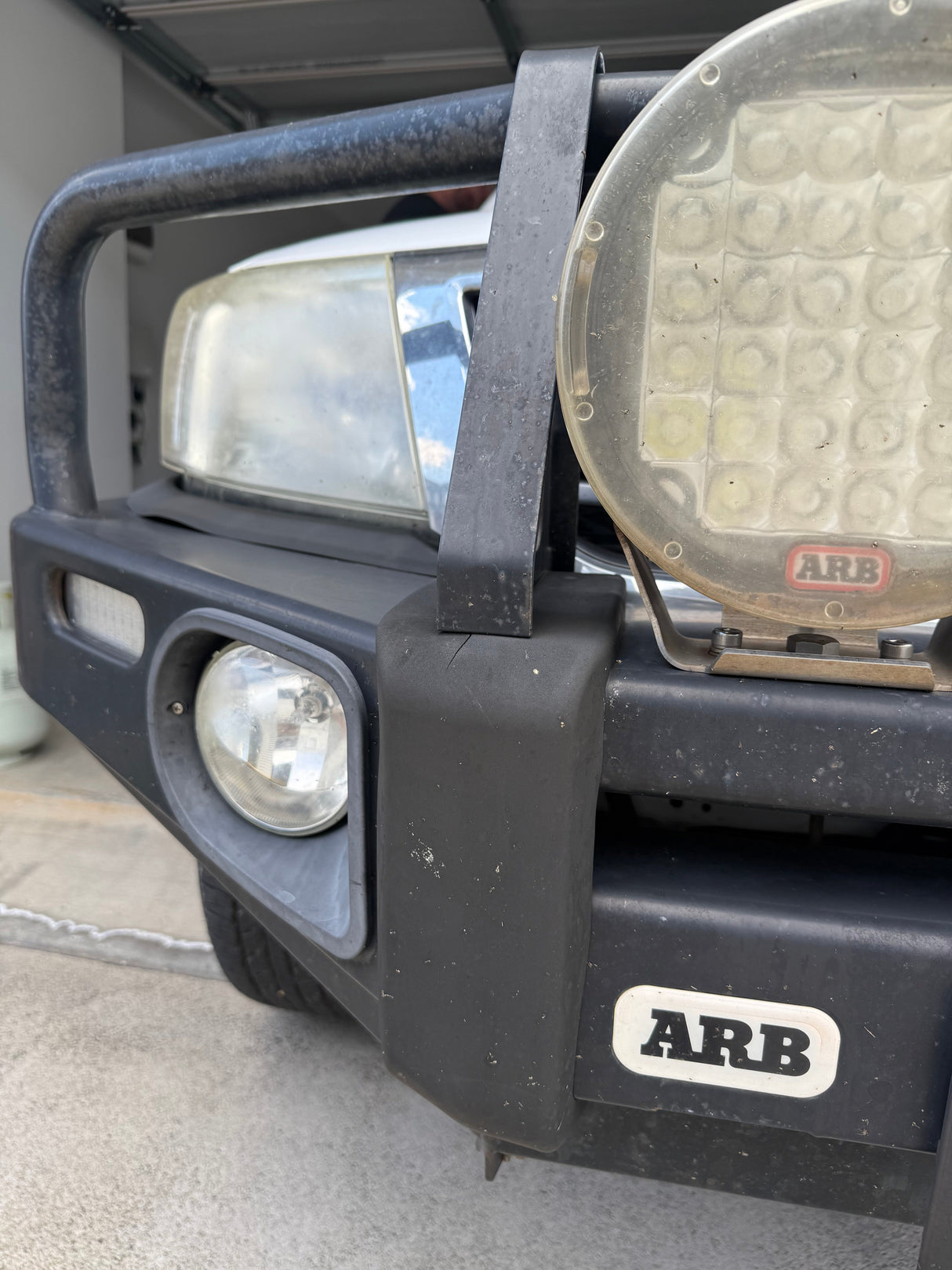 Close-up of a black ARB front bumper and bull bar on an off-road vehicle, showing the main headlight, auxiliary round light, and a white ARB logo badge on the bumper.