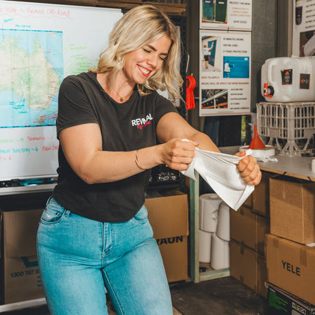 A woman with blonde hair wearing a black t-shirt with 'REVIVAL' printed on the chest and blue jeans, smiling as she holds up a white cloth in a storage room or warehouse setting.