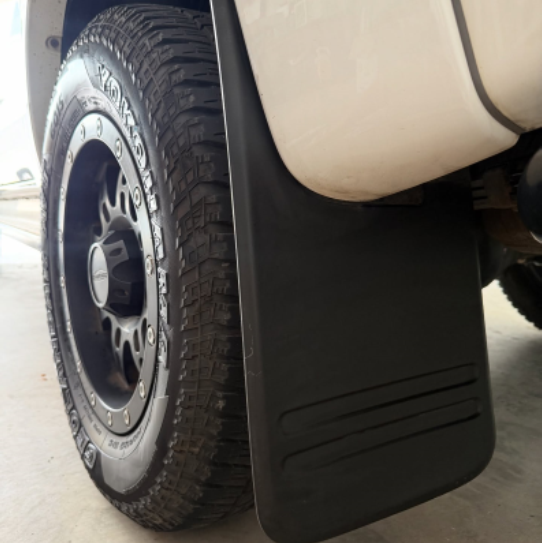 A close-up shot of a black rubber vehicle mud flap mounted on the side of a white truck, with a black tire and off-road tread visible.
