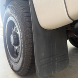 A close-up shot of a black rubber mud flap attached to the side of a white vehicle, with a black off-road tire visible on a concrete floor.