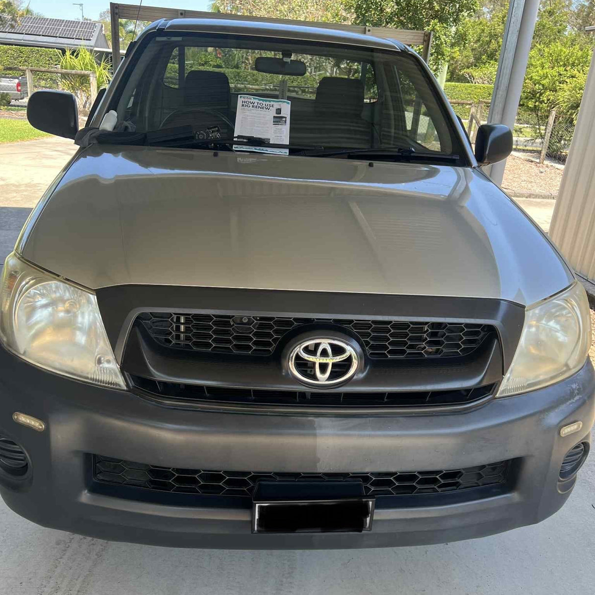 A front-facing silver Toyota pickup truck parked under a carport on a concrete surface, with trees and outdoor structures visible in the background.
