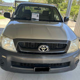 A front-facing silver Toyota pickup truck parked under a carport on a concrete surface, with trees and outdoor structures visible in the background.